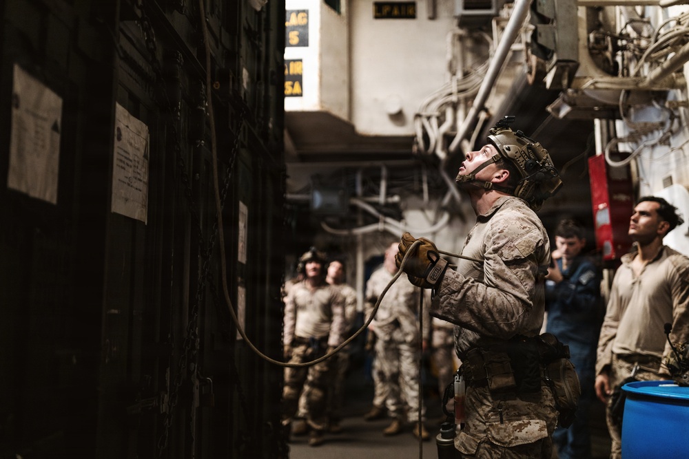 Military personnel, appearing to be U.S. Marines based on their desert camouflage uniforms, are gathered in what appears to be the interior of a naval vessel or military facility. The central figure is wearing a tactical helmet and gloves, holding a rope or cable while looking upward, suggesting rappelling or rope-handling training. Several other personnel in similar uniforms observe in the background.