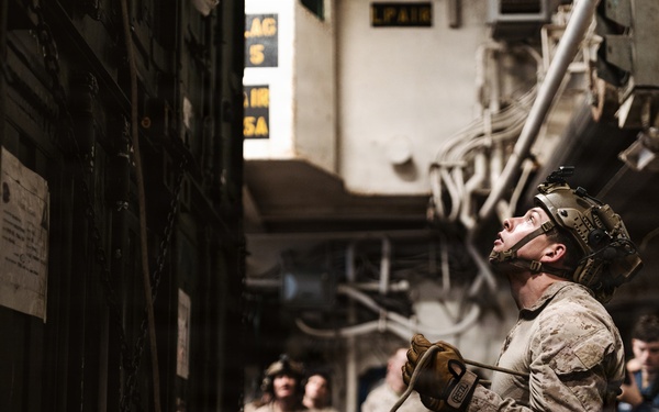 Military personnel, appearing to be U.S. Marines based on their desert camouflage uniforms, are gathered in what appears to be the interior of a naval vessel or military facility. The central figure is wearing a tactical helmet and gloves, holding a rope or cable while looking upward, suggesting rappelling or rope-handling training. Several other personnel in similar uniforms observe in the background.