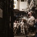 Military personnel, appearing to be U.S. Marines based on their desert camouflage uniforms, are gathered in what appears to be the interior of a naval vessel or military facility. The central figure is wearing a tactical helmet and gloves, holding a rope or cable while looking upward, suggesting rappelling or rope-handling training. Several other personnel in similar uniforms observe in the background.