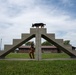 A handler works with a German Shepherd dog on a concrete agility obstacle course at what appears to be a military or law enforcement K9 training facility. The dog is positioned at the apex of a stepped concrete A-frame structure while the handler stands below, looking up and directing the animal. The setting includes institutional buildings, chain-link fencing, and a well-maintained grass field in the background.
