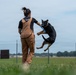 A dog trainer is working with a black and tan German Shepherd in an outdoor training field. The trainer, wearing brown overalls and a black shirt with 'TRAIN' visible on the back, is holding a blue tug toy while the dog leaps into the air to grab it. The setting appears to be a secured facility with chain-link fencing and an open grassy field.