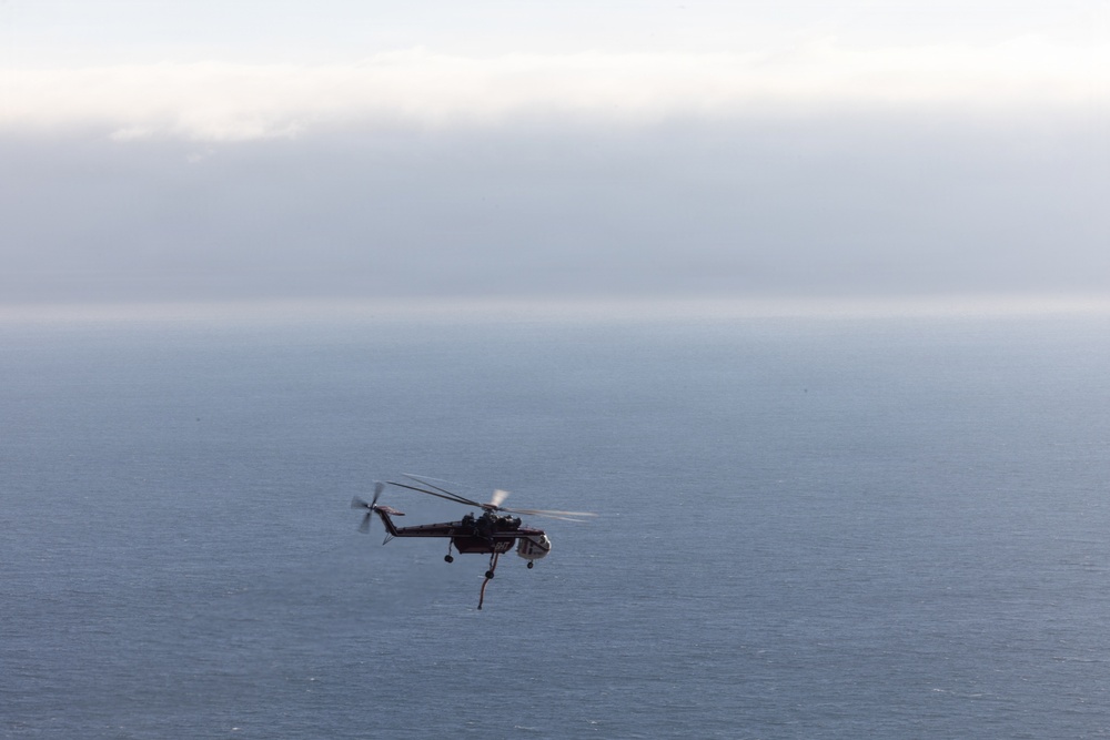 A helicopter is flying over a large body of open water, appearing to carry a suspended load beneath it. The scene is captured against a calm, overcast sky with a misty horizon blending sea and clouds. The helicopter appears to be a heavy-lift type engaged in an external load operation.