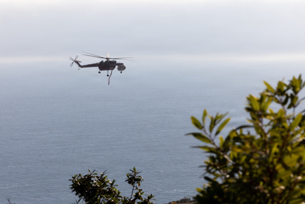 A large helicopter is flying over a coastal ocean scene, photographed from an elevated vantage point with vegetation in the foreground. The helicopter appears to have a sling load or cable hanging beneath it, suggesting it may be engaged in a cargo transport or firefighting water-collection operation. The scene is overcast with hazy atmospheric conditions over the water.