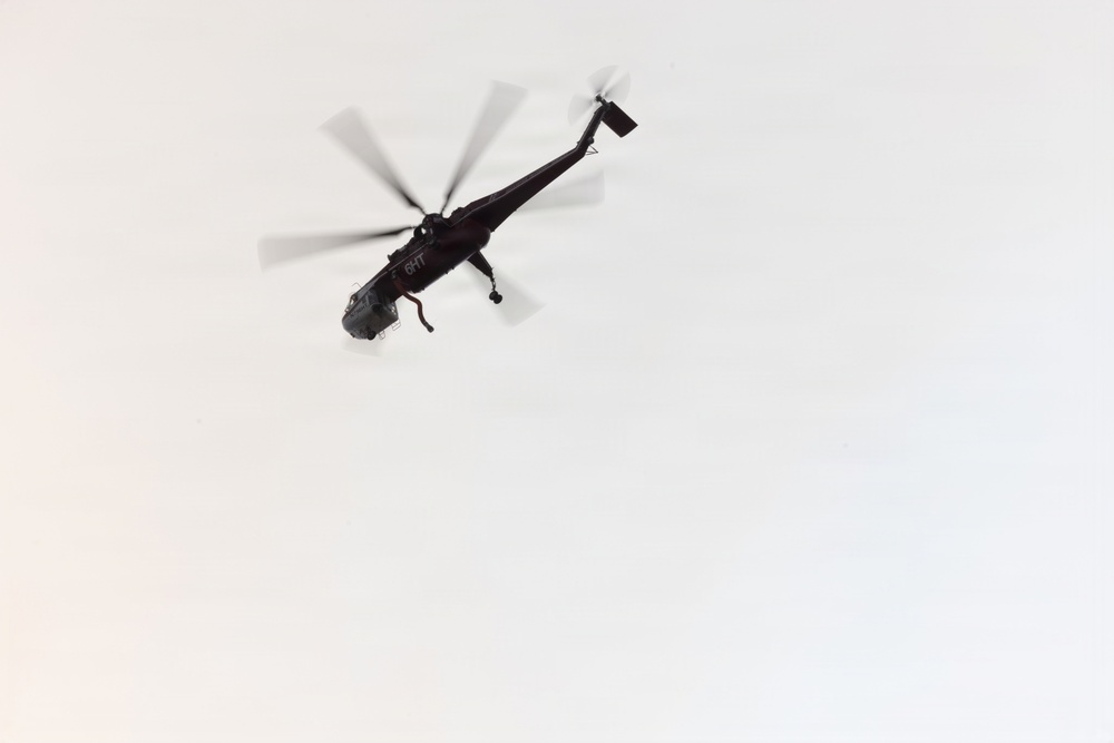A dark-colored helicopter is photographed from below against a pale, overcast sky. The image captures the aircraft in flight with rotor blades in motion, creating a motion blur effect. The helicopter appears to have markings or identifiers visible on its fuselage.