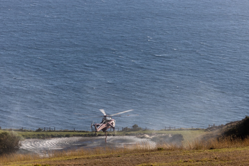 A helicopter is hovering or landing on a coastal clifftop area near the ocean. The rotor wash is visibly disturbing water or wet grass below the aircraft, creating circular ripple patterns on the ground. The helicopter appears to have red and white markings, possibly indicating a rescue or emergency services aircraft.