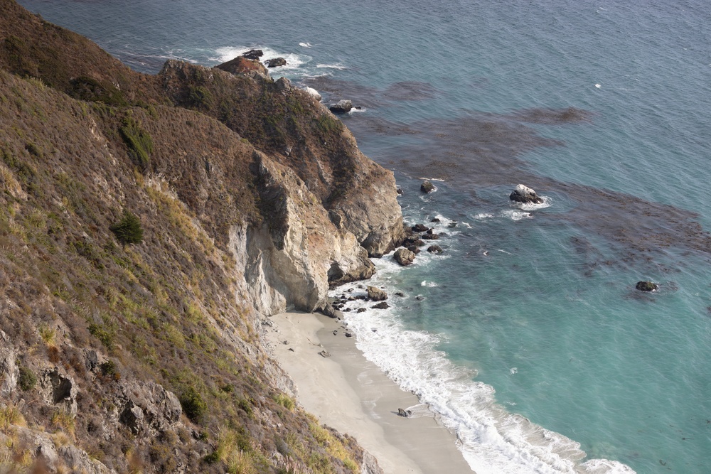 An aerial or elevated view of a rugged coastal cliff meeting a narrow sandy beach along a turquoise ocean. The rocky headland is covered with dry scrub vegetation and features dramatic white and tan limestone cliff faces dropping to the shoreline. Dark kelp beds are visible in the water offshore, and gentle waves wash onto the secluded beach below.