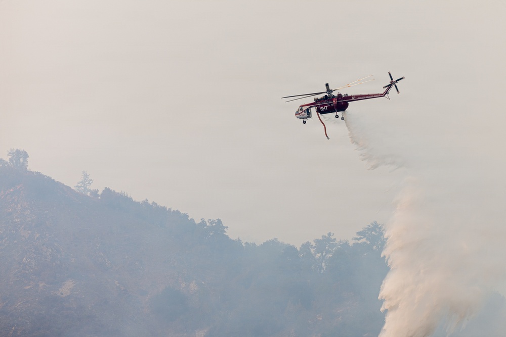 A firefighting helicopter, appearing to be a Sikorsky Skycrane (S-64) in red and white livery, is conducting an aerial water drop operation over a smoke-filled, forested mountainside. The aircraft is actively releasing a large volume of water from its belly tank onto what appears to be an active wildfire below. The scene is heavily obscured by smoke, indicating an ongoing and significant fire event.