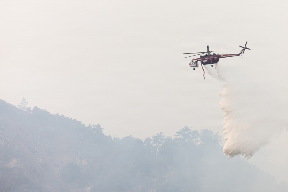 A firefighting helicopter is conducting an aerial water drop over a smoke-filled, forested hillside. The aircraft, marked with what appears to be 'GHT' on its fuselage, is releasing a large cascade of water or fire retardant toward the tree line below. The scene is heavily obscured by smoke, indicating an active wildfire suppression operation.