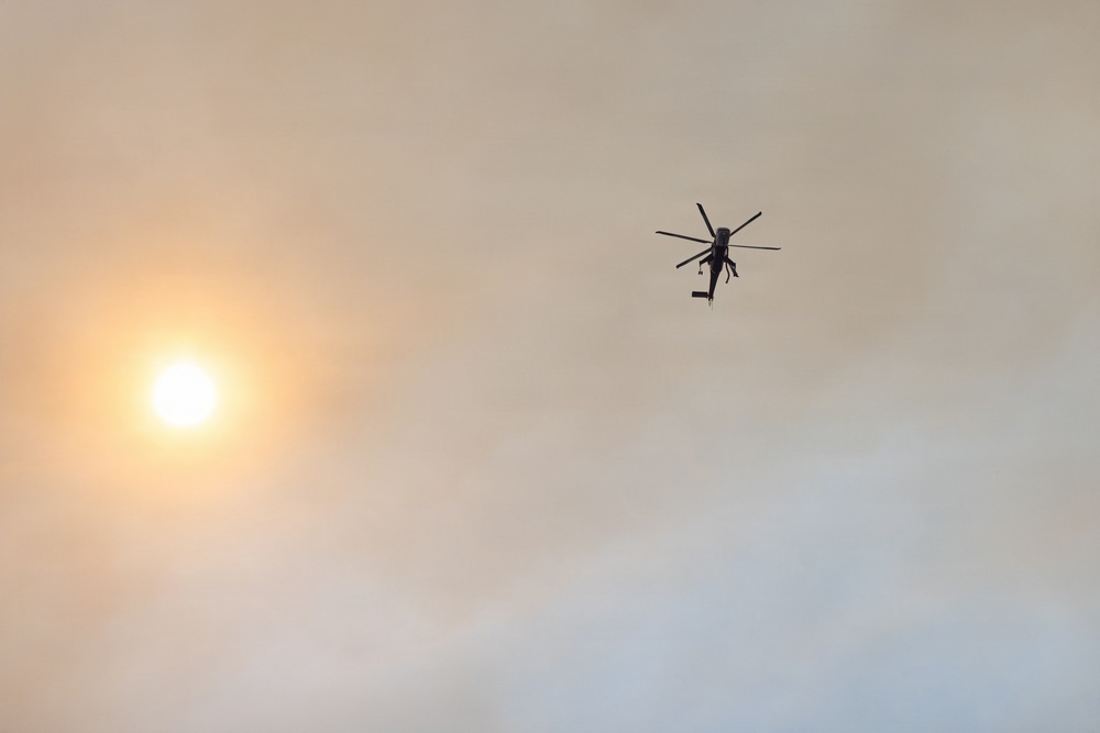 A helicopter is captured in silhouette flying through a hazy, smoke-filled sky. The sun appears as a diffused orange-white orb on the left side of the frame, its light muted by what appears to be smoke or heavy atmospheric haze. The overall scene suggests the helicopter may be operating in conditions related to a wildfire or similar environmental event.