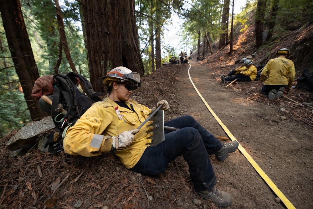 A group of wildland firefighters in yellow Nomex gear are resting along a dirt trail in a dense forest, likely during a break from fire suppression operations. The foreground shows a female firefighter leaning against a tree base, holding a hand tool (appears to be a Pulaski or similar firefighting tool). A yellow hose line runs along the trail connecting the crew members visible in the background.