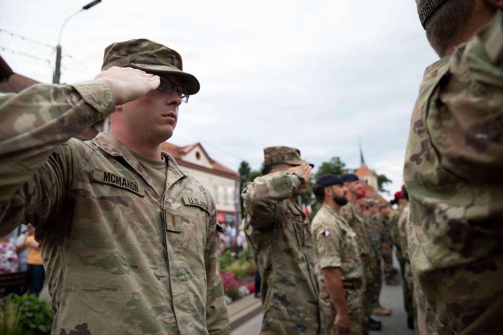 A group of U.S. Army soldiers in MultiCam/OCP camouflage uniforms are performing a salute during what appears to be a formal military ceremony or parade. The soldier in the foreground is identified by his name tape as 'MCMAHON' and wears U.S. Army insignia. The setting appears to be an outdoor public square or street in a European town, with civilian onlookers visible in the background.