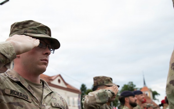 A group of U.S. Army soldiers in MultiCam/OCP camouflage uniforms are performing a salute during what appears to be a formal military ceremony or parade. The soldier in the foreground is identified by his name tape as 'MCMAHON' and wears U.S. Army insignia. The setting appears to be an outdoor public square or street in a European town, with civilian onlookers visible in the background.