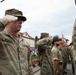 A group of U.S. Army soldiers in MultiCam/OCP camouflage uniforms are performing a salute during what appears to be a formal military ceremony or parade. The soldier in the foreground is identified by his name tape as 'MCMAHON' and wears U.S. Army insignia. The setting appears to be an outdoor public square or street in a European town, with civilian onlookers visible in the background.