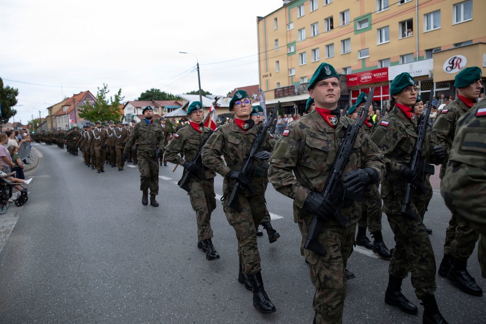 A military parade is taking place on a public street in what appears to be a Polish town, based on visible Polish flag patches on uniforms and Polish-language signage on surrounding buildings. Soldiers in camouflage uniforms, green berets, and red scarves march in formation while carrying rifles. Civilians line the street to observe the parade.