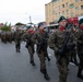 A military parade is taking place on a public street in what appears to be a Polish town, based on visible Polish flag patches on uniforms and Polish-language signage on surrounding buildings. Soldiers in camouflage uniforms, green berets, and red scarves march in formation while carrying rifles. Civilians line the street to observe the parade.