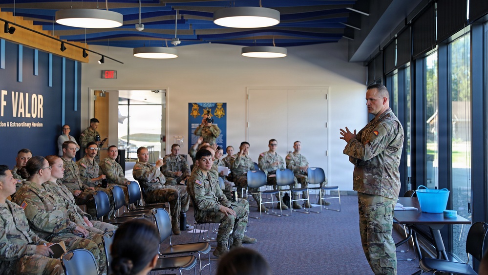 A military briefing or training session is taking place in a modern indoor facility. A single uniformed soldier stands at the front addressing a seated audience of approximately 25-30 military personnel in camouflage uniforms. The setting appears to be a military museum or visitor center, evidenced by signage referencing 'Medal of Valor' and 'Extraordinary Heroism' visible on the wall.