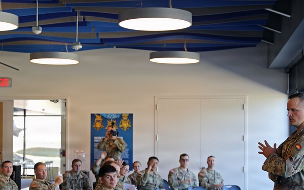 A military briefing or training session is taking place in a modern indoor facility. A single uniformed soldier stands at the front addressing a seated audience of approximately 25-30 military personnel in camouflage uniforms. The setting appears to be a military museum or visitor center, evidenced by signage referencing 'Medal of Valor' and 'Extraordinary Heroism' visible on the wall.