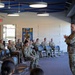 A military briefing or training session is taking place in a modern indoor facility. A single uniformed soldier stands at the front addressing a seated audience of approximately 25-30 military personnel in camouflage uniforms. The setting appears to be a military museum or visitor center, evidenced by signage referencing 'Medal of Valor' and 'Extraordinary Heroism' visible on the wall.