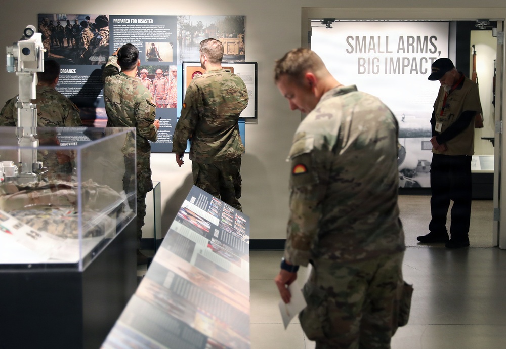 Military personnel in camouflage uniforms are visiting what appears to be a military museum or exhibit hall. Several soldiers are examining display panels and cases, while a civilian staff member stands near a doorway. A prominent exhibit section titled 'Small Arms, Big Impact' is visible in the background.