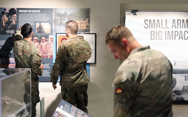Military personnel in camouflage uniforms are visiting what appears to be a military museum or exhibit hall. Several soldiers are examining display panels and cases, while a civilian staff member stands near a doorway. A prominent exhibit section titled 'Small Arms, Big Impact' is visible in the background.
