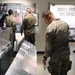 Military personnel in camouflage uniforms are visiting what appears to be a military museum or exhibit hall. Several soldiers are examining display panels and cases, while a civilian staff member stands near a doorway. A prominent exhibit section titled 'Small Arms, Big Impact' is visible in the background.
