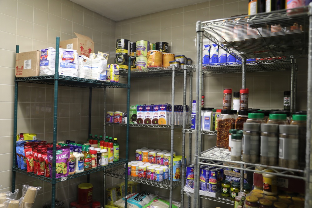 The image shows a well-stocked food pantry or storage room with multiple wire shelving units filled with various dry goods, canned foods, condiments, and spices. The shelves are organized with items grouped by category across several tiers. The room has tiled walls and appears to be a commercial or institutional food storage space.
