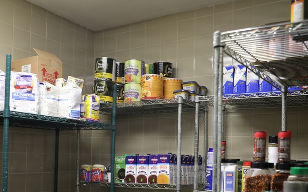 The image shows a well-stocked food pantry or storage room with multiple wire shelving units filled with various dry goods, canned foods, condiments, and spices. The shelves are organized with items grouped by category across several tiers. The room has tiled walls and appears to be a commercial or institutional food storage space.