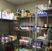 The image shows a well-stocked food pantry or storage room with multiple wire shelving units filled with various dry goods, canned foods, condiments, and spices. The shelves are organized with items grouped by category across several tiers. The room has tiled walls and appears to be a commercial or institutional food storage space.