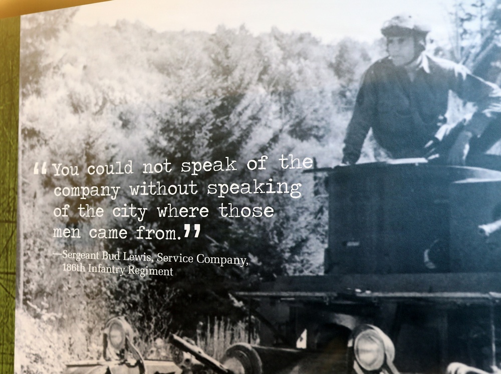 A museum or exhibit display panel featuring a black-and-white historical photograph of a soldier operating what appears to be a military vehicle. Overlaid on the image is a quote attributed to Sergeant Bud Lewis of the Service Company, 186th Infantry Regiment. The quote reads: 'You could not speak of the company without speaking of the city where those men came from.'