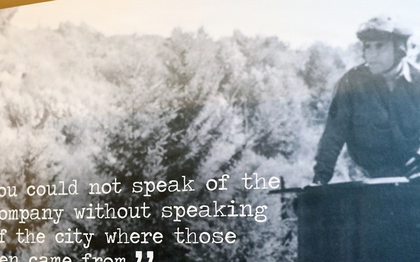 A museum or exhibit display panel featuring a black-and-white historical photograph of a soldier operating what appears to be a military vehicle. Overlaid on the image is a quote attributed to Sergeant Bud Lewis of the Service Company, 186th Infantry Regiment. The quote reads: 'You could not speak of the company without speaking of the city where those men came from.'