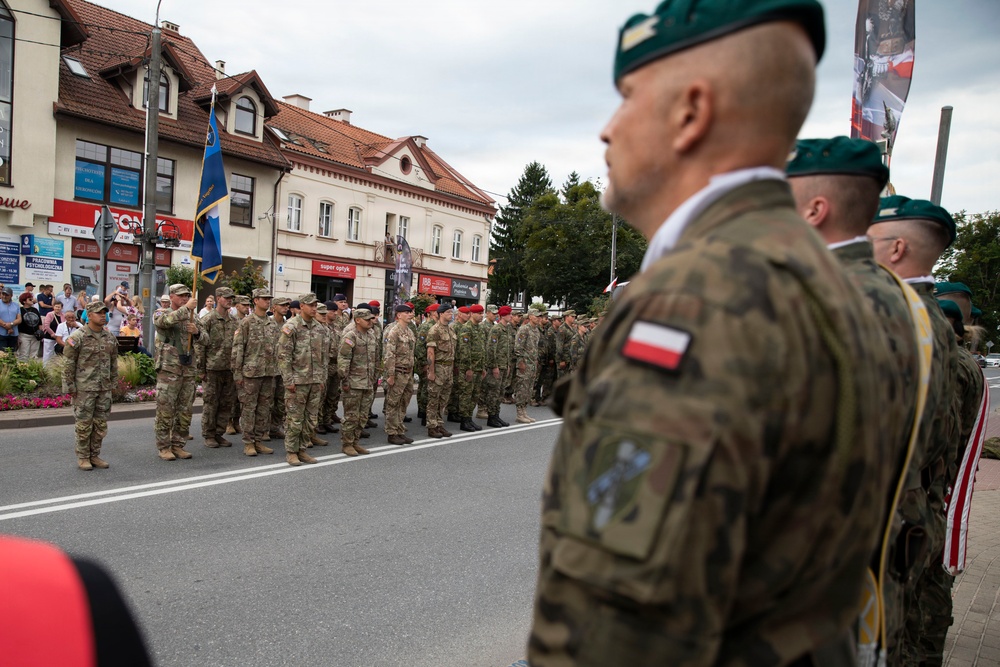 A military ceremony or parade is taking place on a street in what appears to be a Polish town, with soldiers from multiple nations standing in formation. Visible Polish Army soldiers wearing green berets and camouflage uniforms with Polish flag patches are in the foreground, while a formation of soldiers in US-style MultiCam uniforms stands across the street. Civilians and spectators are visible in the background near storefronts.