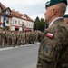 A military ceremony or parade is taking place on a street in what appears to be a Polish town, with soldiers from multiple nations standing in formation. Visible Polish Army soldiers wearing green berets and camouflage uniforms with Polish flag patches are in the foreground, while a formation of soldiers in US-style MultiCam uniforms stands across the street. Civilians and spectators are visible in the background near storefronts.
