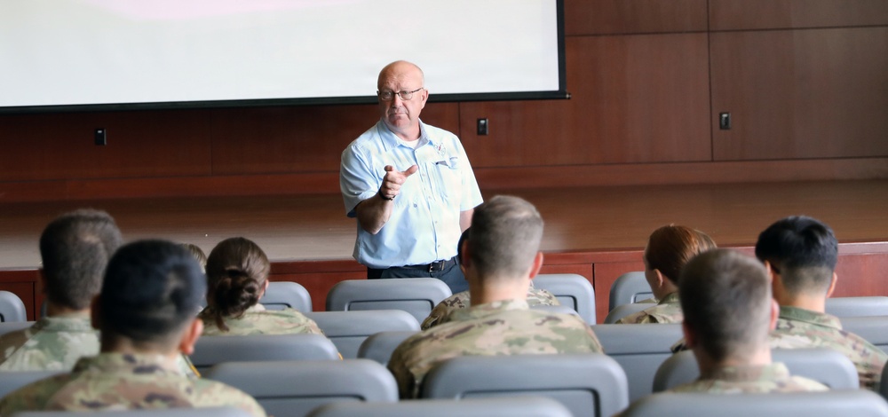 A civilian instructor or speaker addresses a group of military personnel in a briefing room or auditorium. The audience, wearing camouflage uniforms, is seated in rows of gray chairs facing the presenter. A large projection screen is visible behind the speaker, who appears to be gesturing toward the audience.