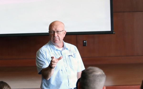 A civilian instructor or speaker addresses a group of military personnel in a briefing room or auditorium. The audience, wearing camouflage uniforms, is seated in rows of gray chairs facing the presenter. A large projection screen is visible behind the speaker, who appears to be gesturing toward the audience.