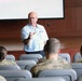 A civilian instructor or speaker addresses a group of military personnel in a briefing room or auditorium. The audience, wearing camouflage uniforms, is seated in rows of gray chairs facing the presenter. A large projection screen is visible behind the speaker, who appears to be gesturing toward the audience.