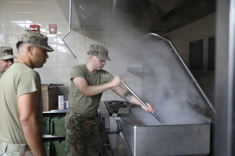 Military personnel in camouflage uniforms and olive drab t-shirts are working in a commercial kitchen facility. One soldier is lifting the large lid of an industrial steam kettle or tilting skillet, releasing a significant cloud of steam. The setting appears to be a military dining facility (DFAC) or mess hall.