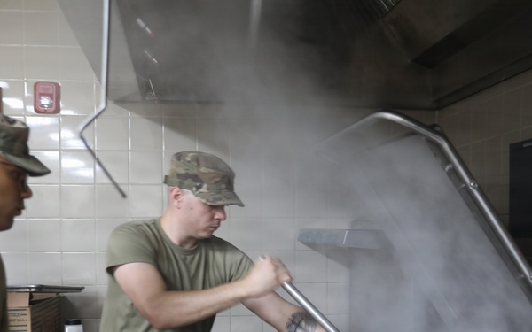 Military personnel in camouflage uniforms and olive drab t-shirts are working in a commercial kitchen facility. One soldier is lifting the large lid of an industrial steam kettle or tilting skillet, releasing a significant cloud of steam. The setting appears to be a military dining facility (DFAC) or mess hall.