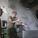Military personnel in camouflage uniforms and olive drab t-shirts are working in a commercial kitchen facility. One soldier is lifting the large lid of an industrial steam kettle or tilting skillet, releasing a significant cloud of steam. The setting appears to be a military dining facility (DFAC) or mess hall.