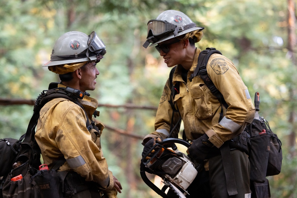 Two wildland firefighters in full gear are engaged in conversation in a forested setting. Both wear standard yellow Nomex fire-resistant shirts, gray helmets with goggles, and dark pants. One firefighter is holding a chainsaw, suggesting active fire line or fuel reduction work.