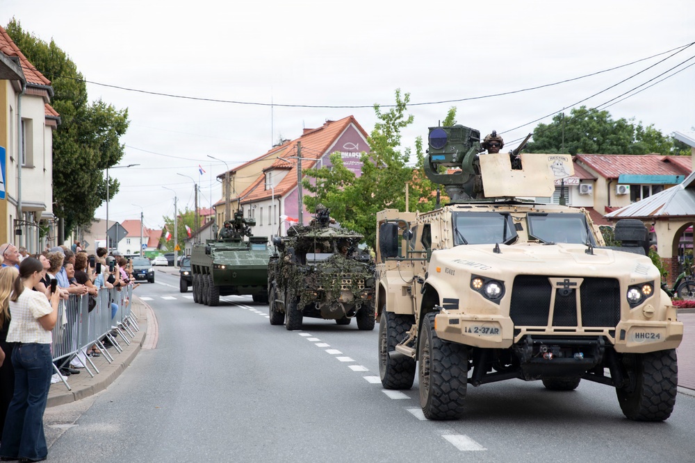 A military convoy parade moves through a small European town street, with civilians watching from behind metal barriers on the left sidewalk. The lead vehicle is a tan/beige JLTV (Joint Light Tactical Vehicle) marked 'HHC 2-4' and '1A 2-37 AR', followed by camouflage-netted wheeled armored vehicles. The setting appears to be a Polish town based on visible Polish flags and signage.