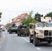 A military convoy parade moves through a small European town street, with civilians watching from behind metal barriers on the left sidewalk. The lead vehicle is a tan/beige JLTV (Joint Light Tactical Vehicle) marked 'HHC 2-4' and '1A 2-37 AR', followed by camouflage-netted wheeled armored vehicles. The setting appears to be a Polish town based on visible Polish flags and signage.