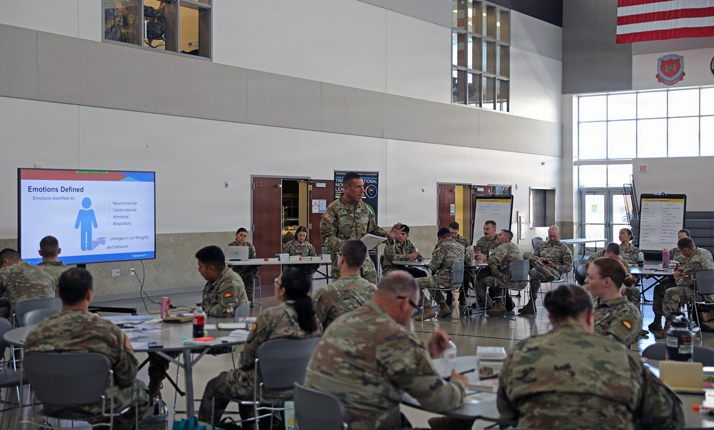 A military training or leadership seminar is taking place in a large indoor facility, with uniformed soldiers seated at round tables facing a presenter. A PowerPoint slide titled 'Emotions Defined' is displayed on a large screen to the left, suggesting a resilience, mental health, or leadership development training session. An American flag and what appears to be a unit insignia are visible on the upper right wall.