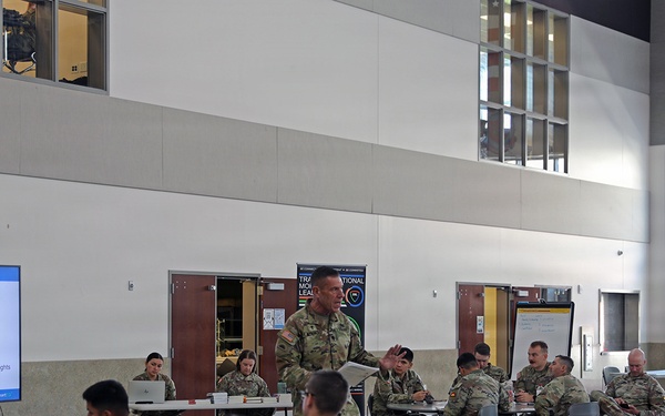 A military training or leadership seminar is taking place in a large indoor facility, with uniformed soldiers seated at round tables facing a presenter. A PowerPoint slide titled 'Emotions Defined' is displayed on a large screen to the left, suggesting a resilience, mental health, or leadership development training session. An American flag and what appears to be a unit insignia are visible on the upper right wall.