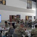 A military training or leadership seminar is taking place in a large indoor facility, with uniformed soldiers seated at round tables facing a presenter. A PowerPoint slide titled 'Emotions Defined' is displayed on a large screen to the left, suggesting a resilience, mental health, or leadership development training session. An American flag and what appears to be a unit insignia are visible on the upper right wall.