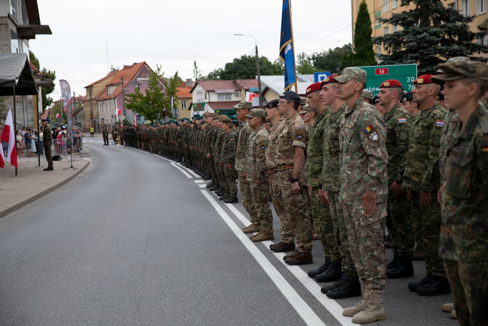 A multinational military formation is standing at attention along a street during what appears to be a formal parade or ceremony. Soldiers wearing various national uniforms and insignia are lined up in a single extended rank, with spectators visible on the left side behind barriers. A blue flag or standard is being carried near the center of the formation.