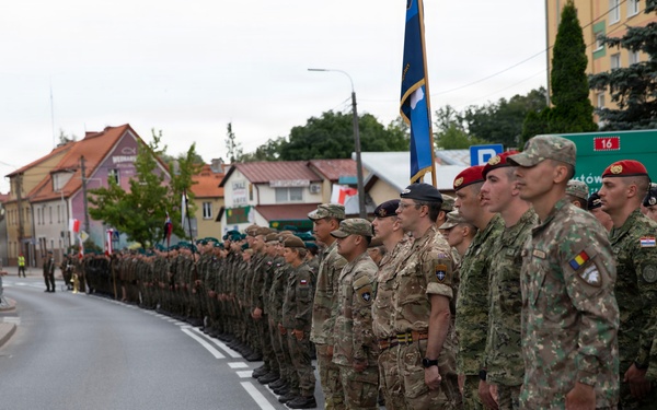 A multinational military formation is standing at attention along a street during what appears to be a formal parade or ceremony. Soldiers wearing various national uniforms and insignia are lined up in a single extended rank, with spectators visible on the left side behind barriers. A blue flag or standard is being carried near the center of the formation.