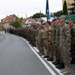 A multinational military formation is standing at attention along a street during what appears to be a formal parade or ceremony. Soldiers wearing various national uniforms and insignia are lined up in a single extended rank, with spectators visible on the left side behind barriers. A blue flag or standard is being carried near the center of the formation.
