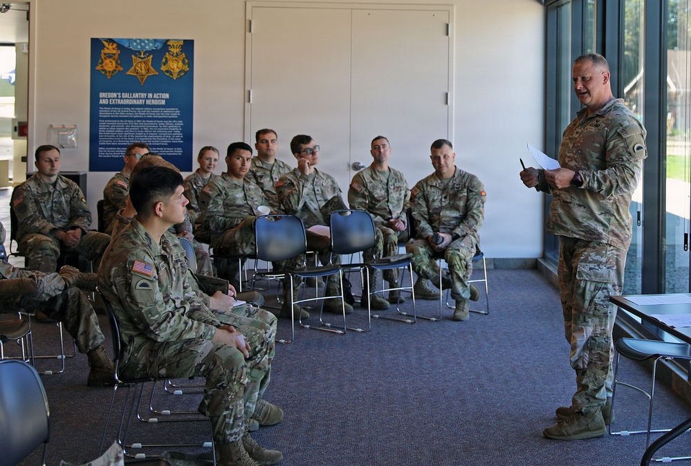 A U.S. Army military briefing or training session is taking place indoors, with one senior soldier standing and addressing a group of approximately 12-15 seated soldiers in military camouflage uniforms. The setting appears to be a visitor center or museum space, as evidenced by a display poster on the wall titled 'Oregon's Gallantry in Action and Extraordinary Heroism' featuring Medal of Honor imagery. The standing soldier holds papers and appears to be conducting an official presentation or instruction.