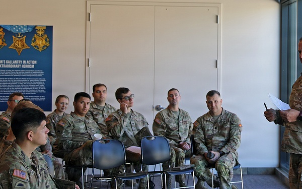 A U.S. Army military briefing or training session is taking place indoors, with one senior soldier standing and addressing a group of approximately 12-15 seated soldiers in military camouflage uniforms. The setting appears to be a visitor center or museum space, as evidenced by a display poster on the wall titled 'Oregon's Gallantry in Action and Extraordinary Heroism' featuring Medal of Honor imagery. The standing soldier holds papers and appears to be conducting an official presentation or instruction.