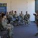 A U.S. Army military briefing or training session is taking place indoors, with one senior soldier standing and addressing a group of approximately 12-15 seated soldiers in military camouflage uniforms. The setting appears to be a visitor center or museum space, as evidenced by a display poster on the wall titled 'Oregon's Gallantry in Action and Extraordinary Heroism' featuring Medal of Honor imagery. The standing soldier holds papers and appears to be conducting an official presentation or instruction.